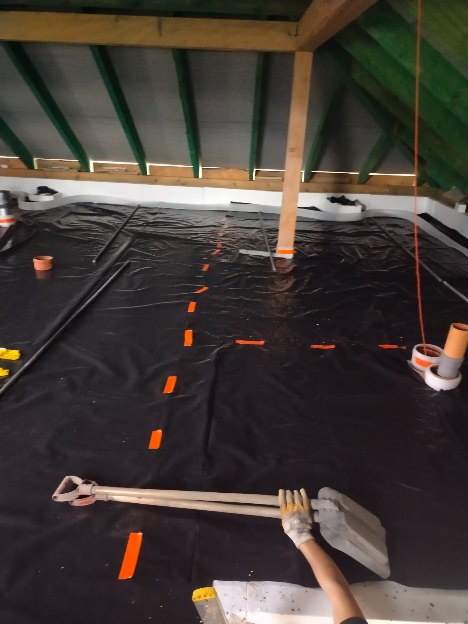 Przygotowanie podłoża pod wylewkę: czarna folia, rury, pomarańczowe znaczniki i narzędzia na poddaszu z drewnianą konstrukcją dachu.