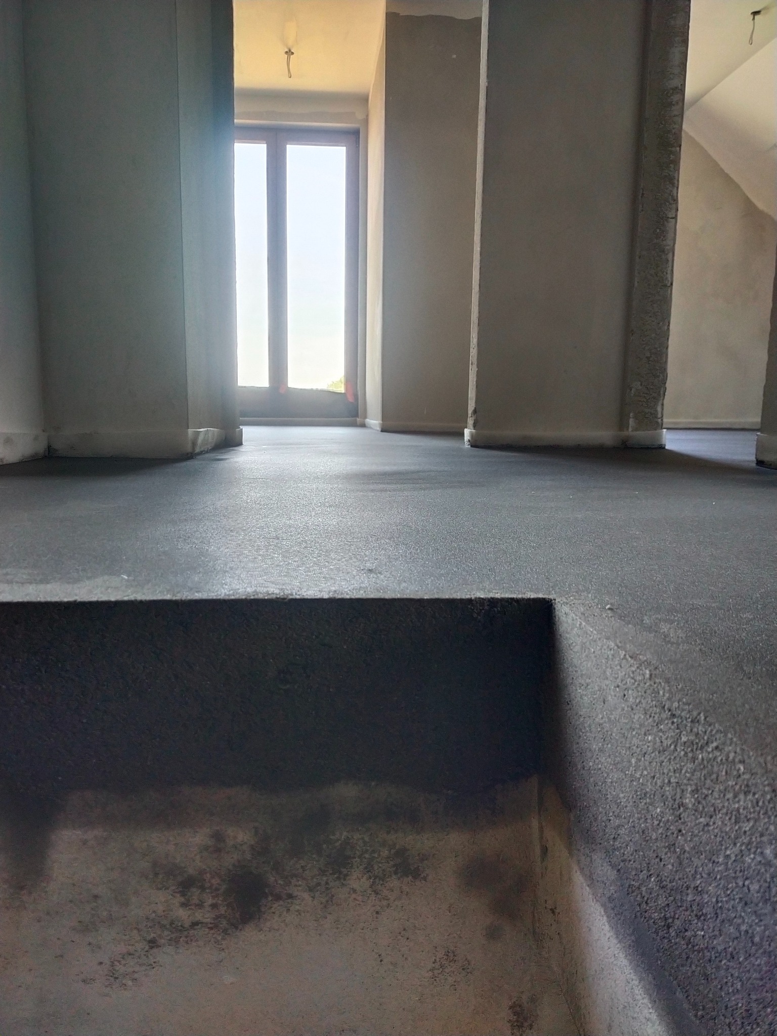 Świeżo wylana, ciemna posadzka w surowym wnętrzu z kolumnami i oknem w tle. Widoczny fragment schodka/podestu. Minimalistyczny, industrialny charakter.