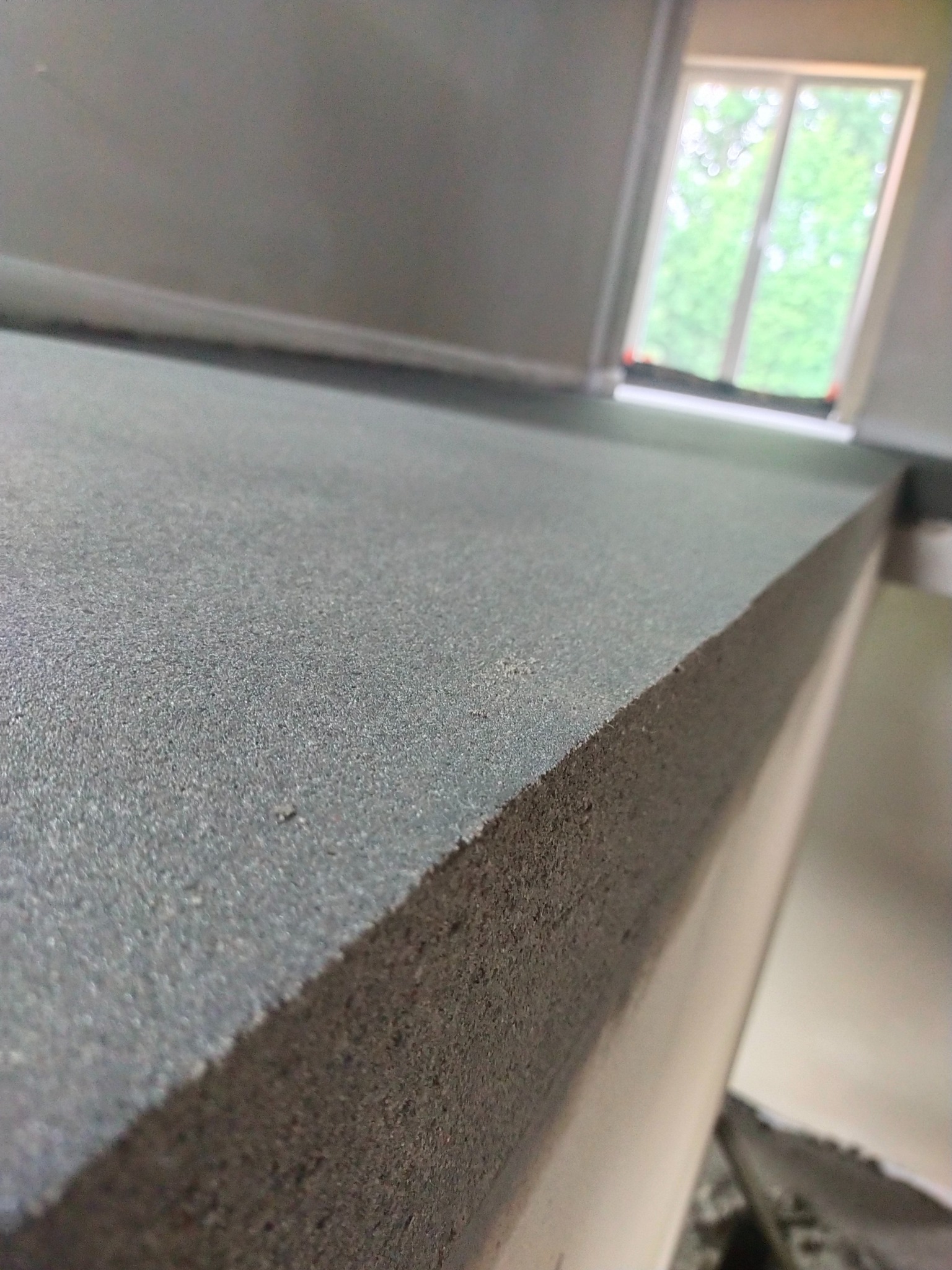 Świeżo wylana, gładka posadzka w szarym odcieniu, widoczna z bliska, z oknem w tle. Ukazany przekrój wylewki.