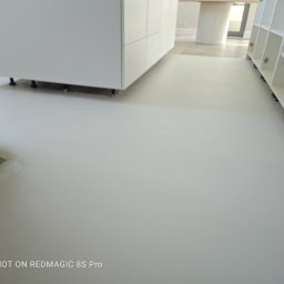 Petro Tychkovskyi - Jasnoszara, gładka posadzka mikrocementowa w minimalistycznym wnętrzu z białymi meblami kuchennymi i regałami. Widoczny fragment podłogi przed wykończeniem.