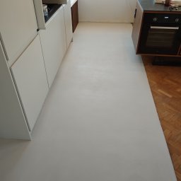 Petro Tychkovskyi - Jasnoszara, gładka posadzka w kuchni, kontrastująca z fragmentem parkietu. Minimalistyczne białe szafki kuchenne i sprzęt AGD w tle. Widok na okno z naturalnym światłem.
