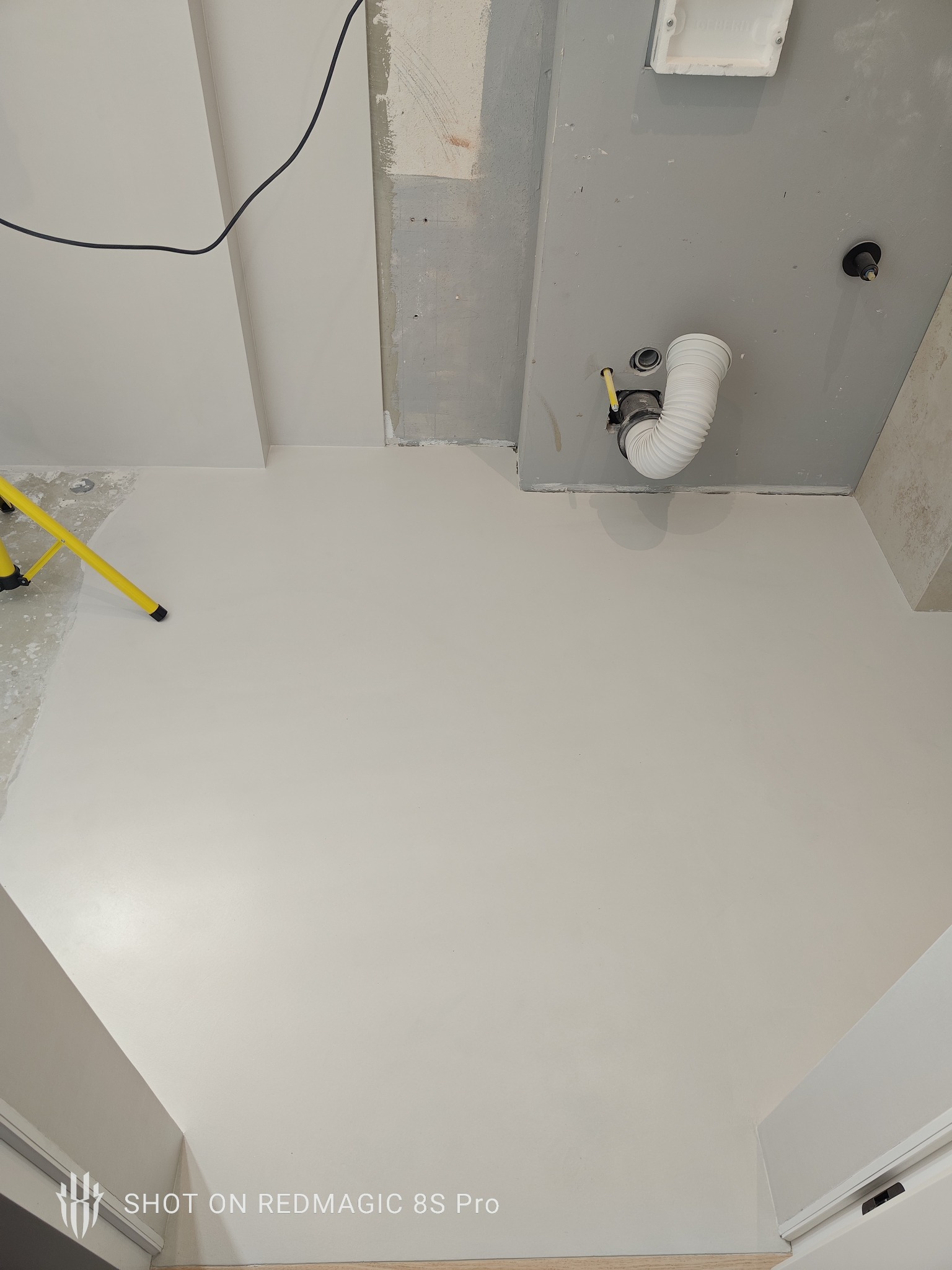 Jasna, gładka posadzka w trakcie remontu łazienki, widoczna drabina malarska i fragment instalacji pod umywalką. Minimalistyczne wykończenie, surowy stan ścian.