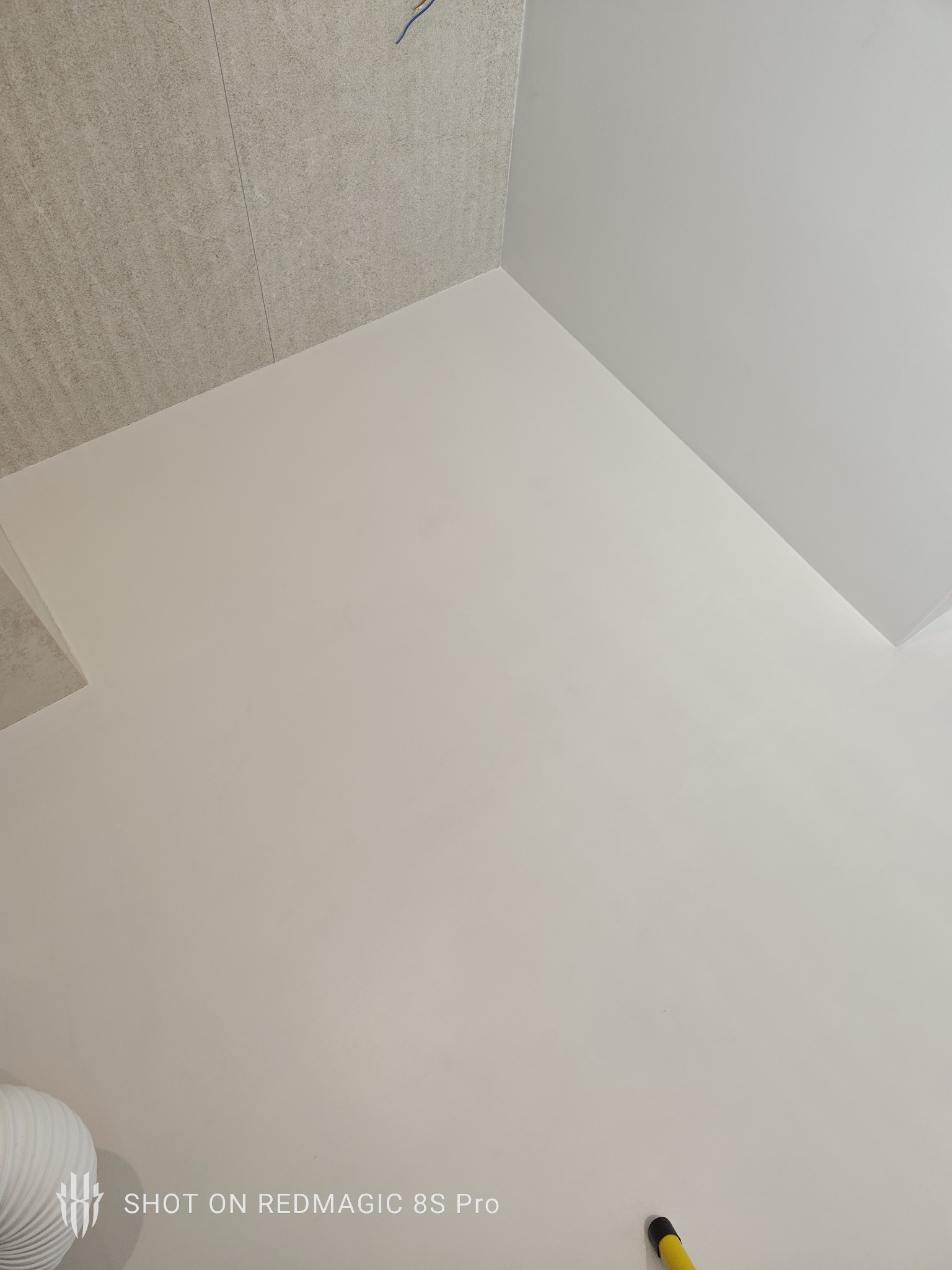 Jasna, gładka posadzka w rogu pomieszczenia, z fragmentem ściany wykończonej płytkami imitującymi kamień. Widoczny fragment rury i narzędzia.