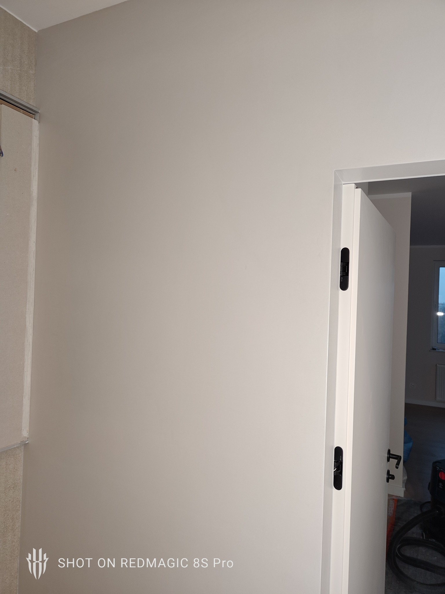 Gładka, beżowa ściana po malowaniu, widoczna z fragmentem białych drzwi z czarnymi klamkami i wykończonym narożnikiem. W tle widoczne wnętrze mieszkania.