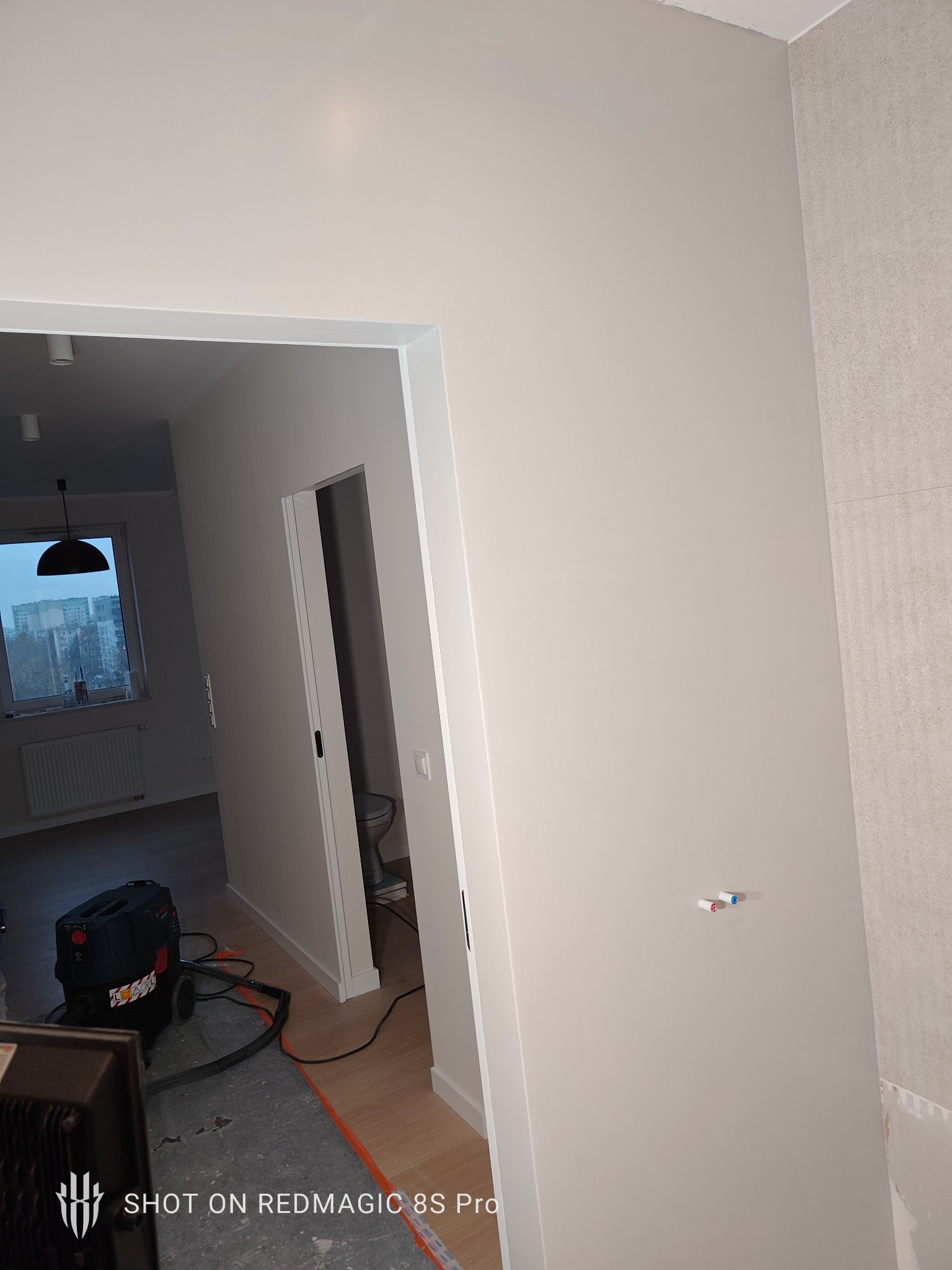 Jasne, gładkie ściany w odcieniach bieli w nowym mieszkaniu, widoczne przyłącza wodne i odkurzacz przemysłowy. Fragment wykończonej łazienki z płytkami w tle.