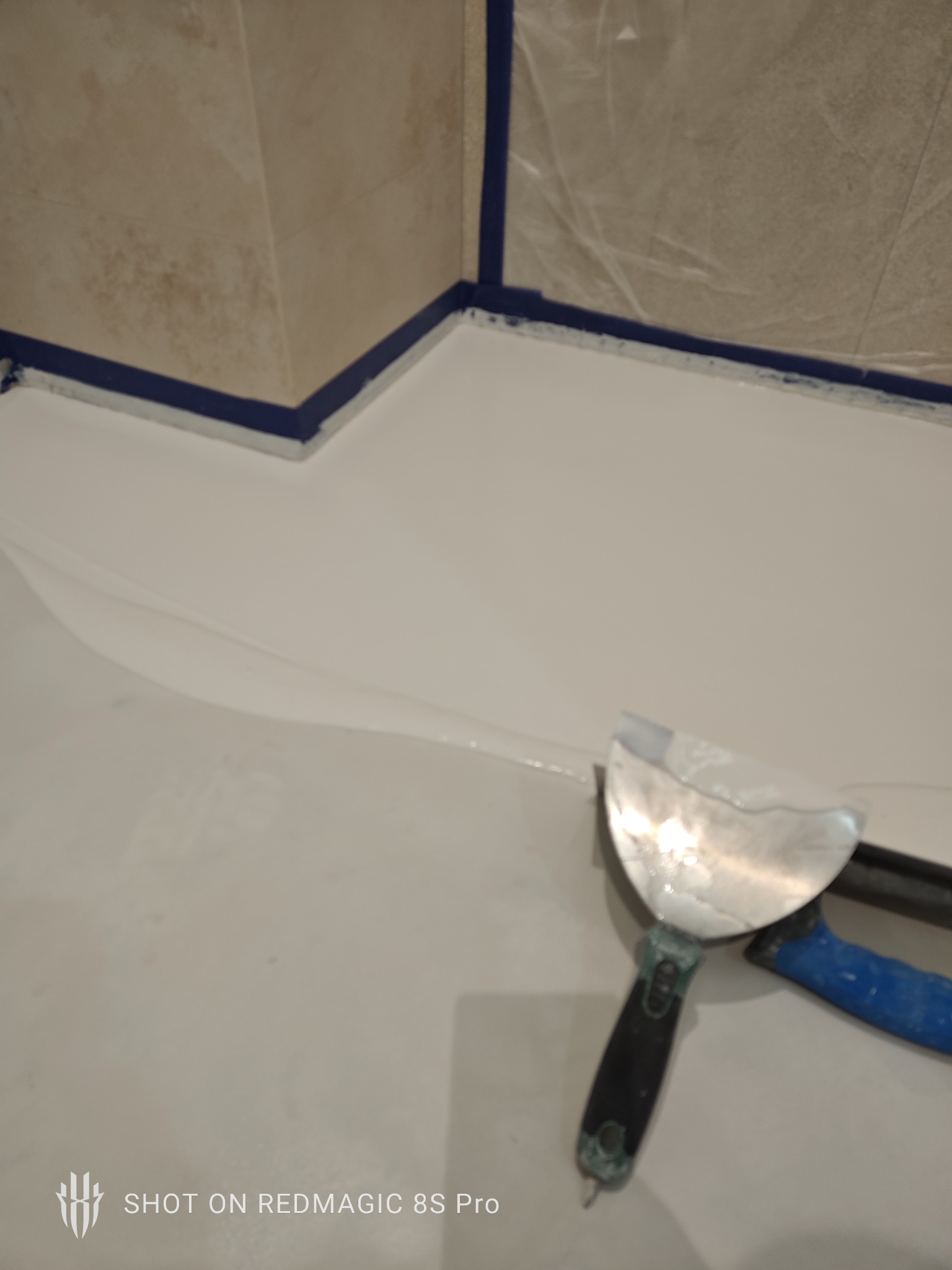 Biała, gładka powierzchnia w rogu pomieszczenia, zabezpieczona niebieską taśmą malarską, z widoczną szpachelką z czarno-niebieską rączką. Przygotowanie do prac wykończeniowych.