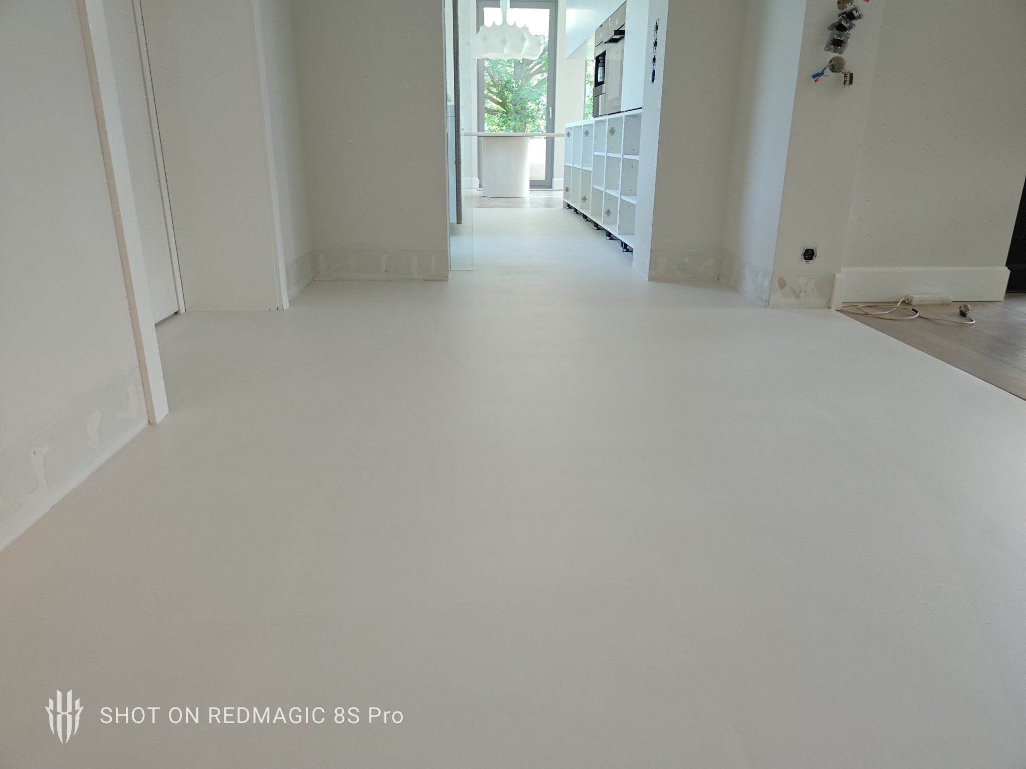 Jasna, jednolita posadzka w przestronnym wnętrzu z białymi ścianami i widocznymi instalacjami elektrycznymi. Minimalistyczny design z otwartą przestrzenią.