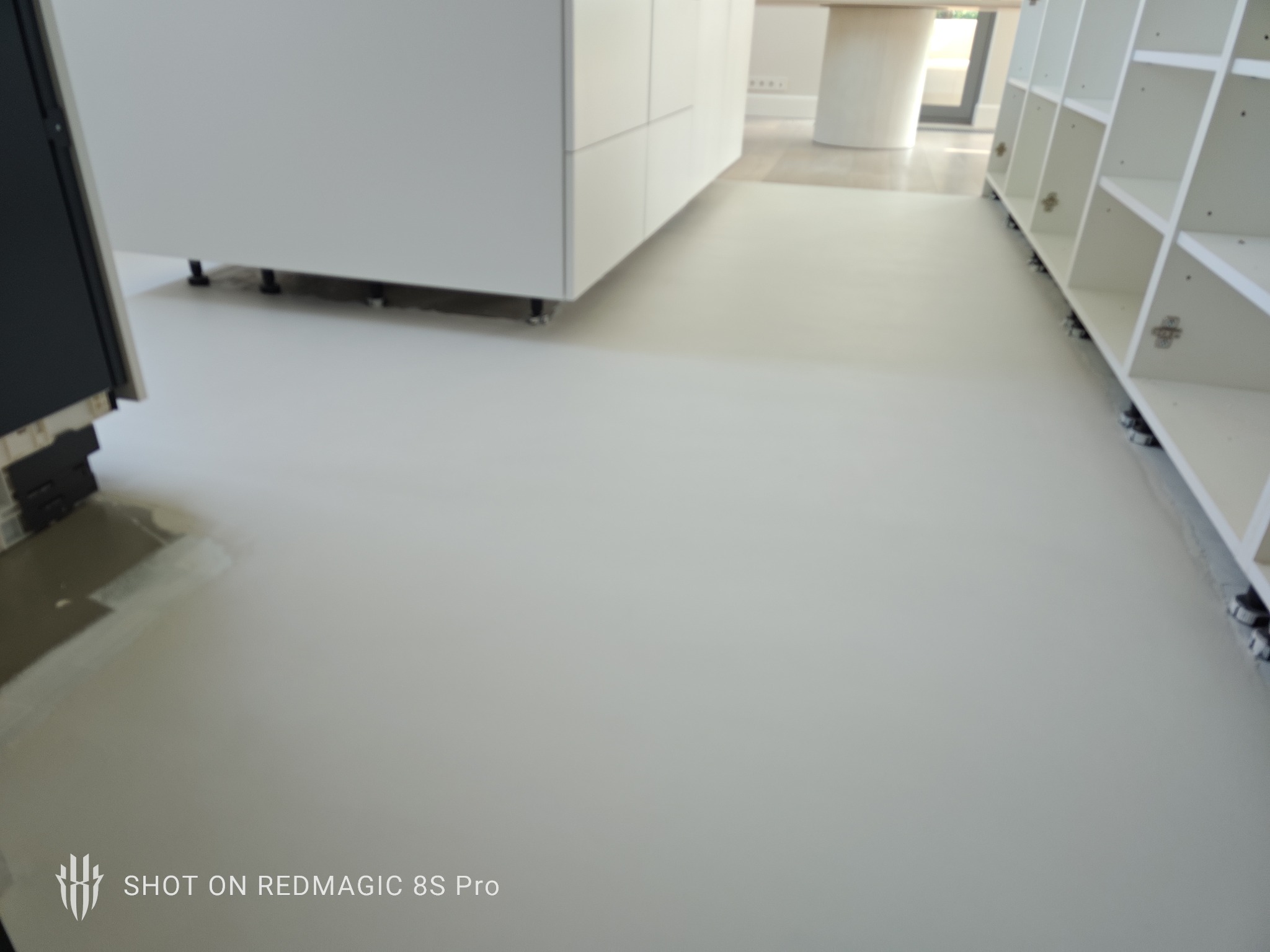 Jasnoszara, gładka posadzka mikrocementowa w minimalistycznym wnętrzu z białymi meblami kuchennymi i regałami. Widoczny fragment podłogi przed wykończeniem.