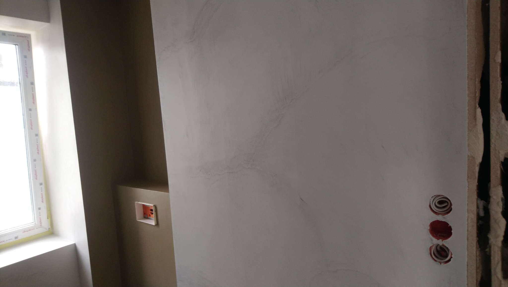 Ściana z efektem dekoracyjnym w odcieniach szarości, widoczne gniazdka elektryczne i fragment okna z parapetem. Minimalistyczny design wnętrza w trakcie wykańczania.