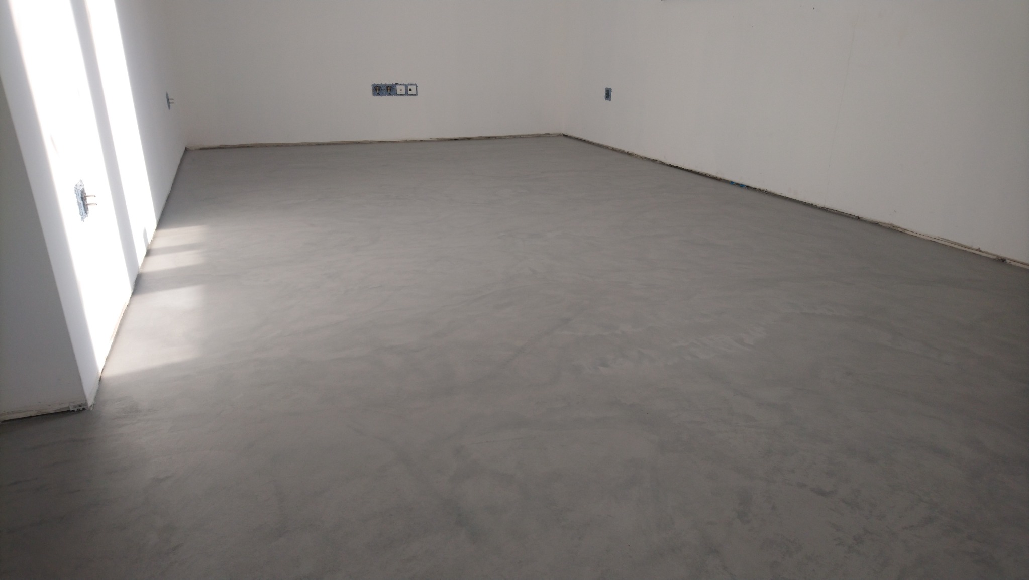 Szara, gładka posadzka mikrocementowa w jasnym pomieszczeniu z białymi ścianami i widocznymi gniazdkami elektrycznymi. Minimalistyczny design.