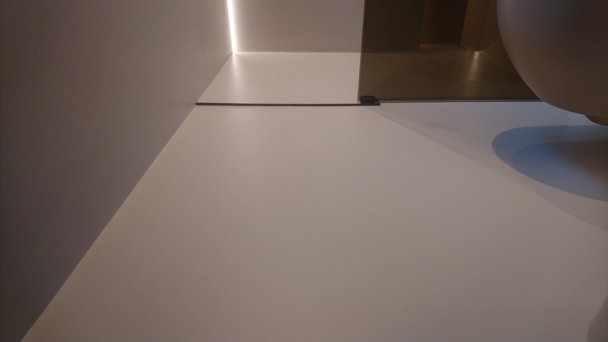 Minimalistyczna łazienka z białymi ścianami, szklanym panelem prysznicowym i subtelnym oświetleniem LED. Widoczny fragment umywalki.