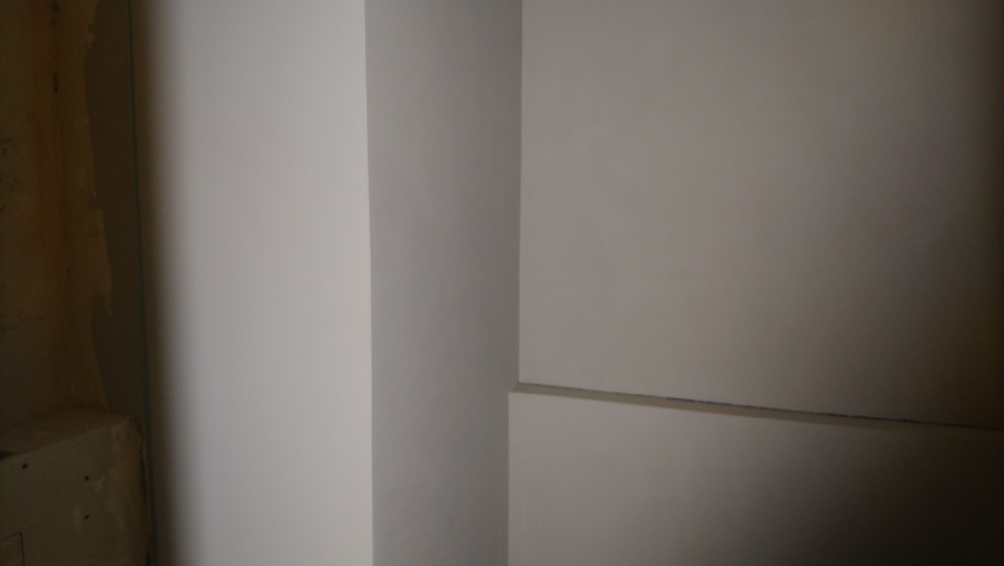 Świeżo pomalowany narożnik pokoju w odcieniach bieli i szarości, widoczne ślady po taśmie malarskiej, surowe wykończenie w tle.