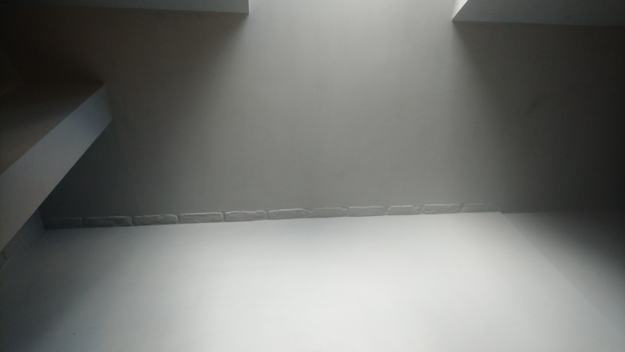 Minimalistyczne wnętrze z białymi ścianami i dekoracyjnym pasem cegieł pod sufitem, oświetlone naturalnym światłem wpadającym z góry. Surowy, nowoczesny design.