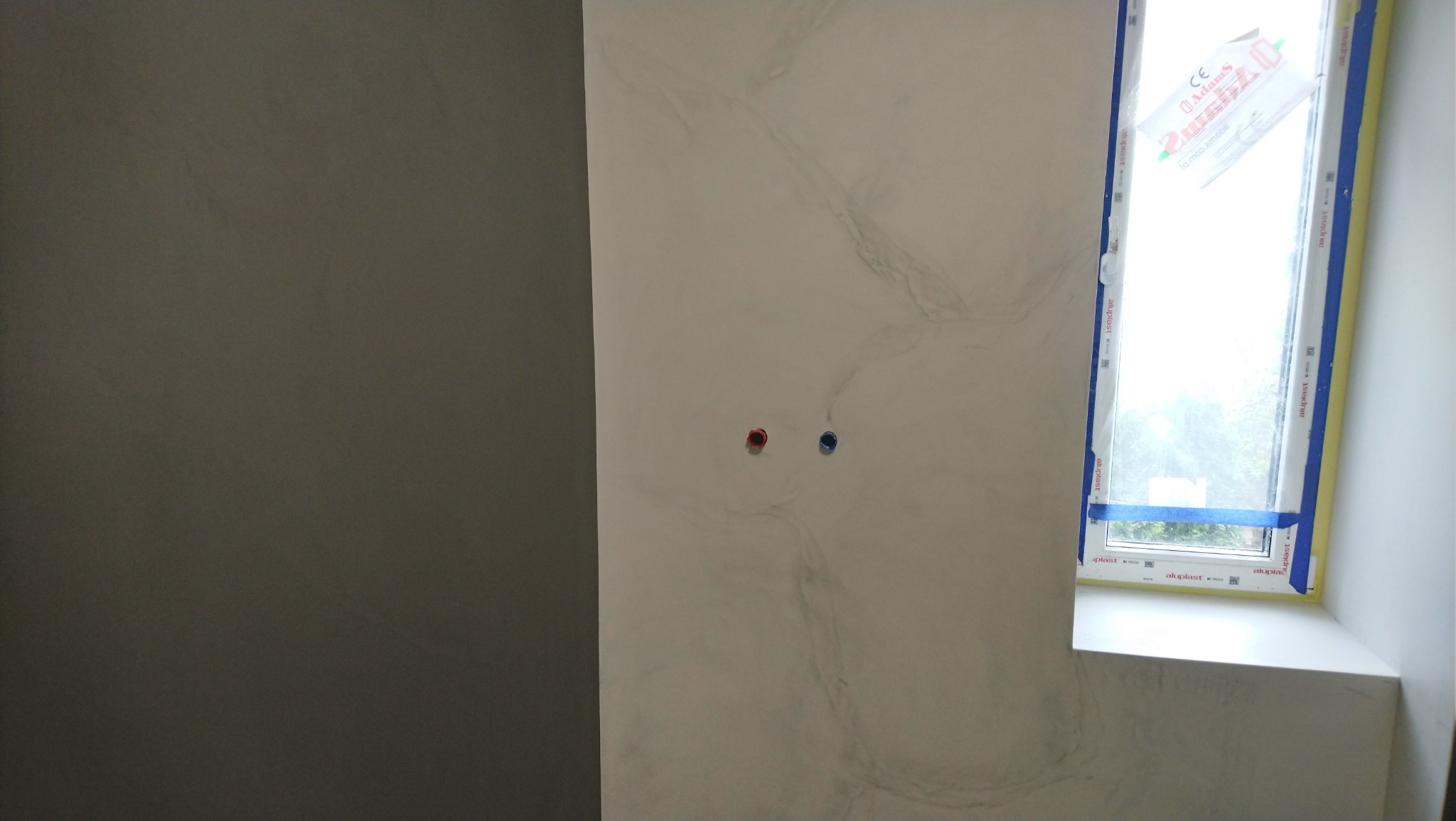 Ściana z efektem marmuru, widoczne gniazdka elektryczne i okno z niebieską taśmą zabezpieczającą, świeżo po wykończeniu.