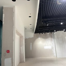 JAKBUD Usługi Budowlane Jakub Soszka - Wnętrze biura w trakcie prac wykończeniowych: ściany z płyt karton-gips, częściowo wyszpachlowane, sufit z kratką i oświetleniem, drabina aluminiowa z boku.