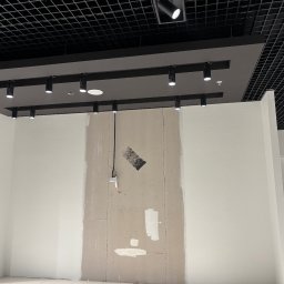 JAKBUD Usługi Budowlane Jakub Soszka - Ściana w trakcie szpachlowania w biurze, widoczne łączenia płyt karton-gips, instalacja elektryczna i czarna kratownica sufitowa z oświetleniem punktowym.