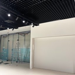 JAKBUD Usługi Budowlane Jakub Soszka - Wnętrze biura w trakcie wykańczania: białe ściany, aluminiowa drabina, przeszklona witryna zabezpieczona folią, czarna kratownica sufitowa z oświetleniem punktowym.