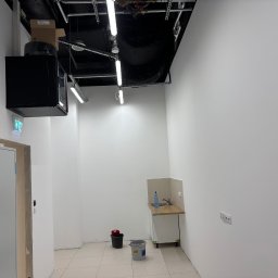 JAKBUD Usługi Budowlane Jakub Soszka - Białe, puste pomieszczenie biurowe po remoncie, z umywalką w rogu, widoczne instalacje pod sufitem i skrzynka elektryczna przy wejściu. Podłoga wyłożona płytkami.