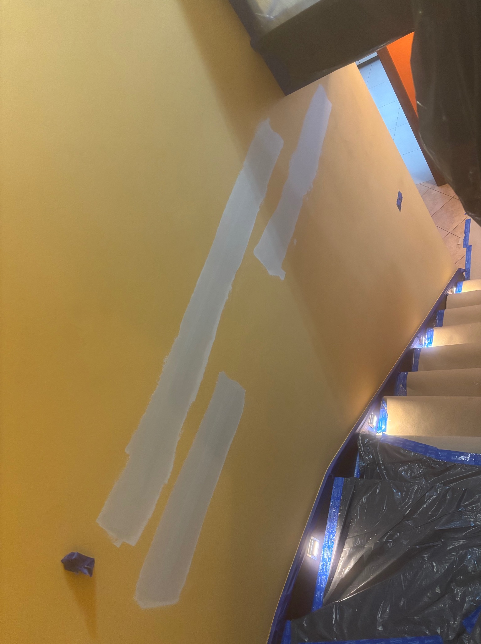 Żółta ściana klatki schodowej w trakcie szpachlowania, zabezpieczona folią i taśmą malarską. Widoczne ślady szpachli i fragment schodów.