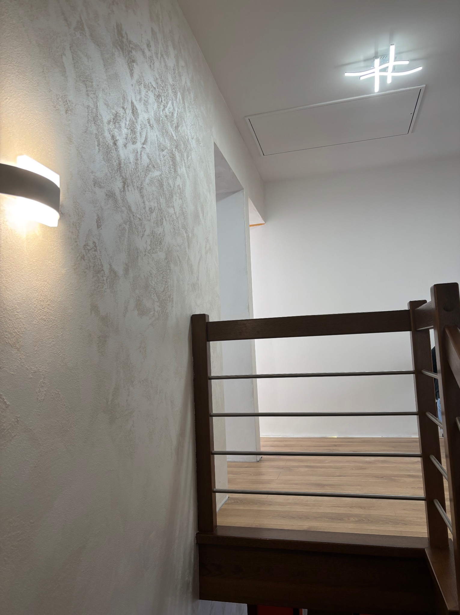 Ściana z dekoracyjną fakturą w odcieniach beżu, oświetlona kinkietem. Widoczne drewniane schody z metalowymi poręczami i fragment sufitu z nowoczesną lampą.