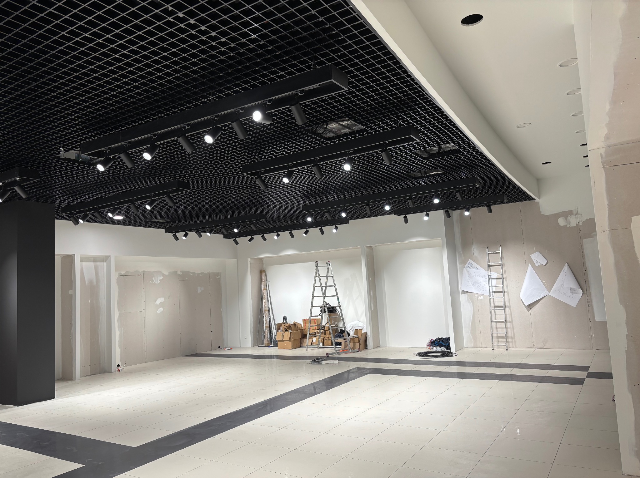 Wnętrze w trakcie wykańczania: sufit z czarną kratownicą i oświetleniem punktowym, ściany częściowo z płyt karton-gips, podłoga z jasnych płytek. Widoczne drabiny i materiały budowlane.