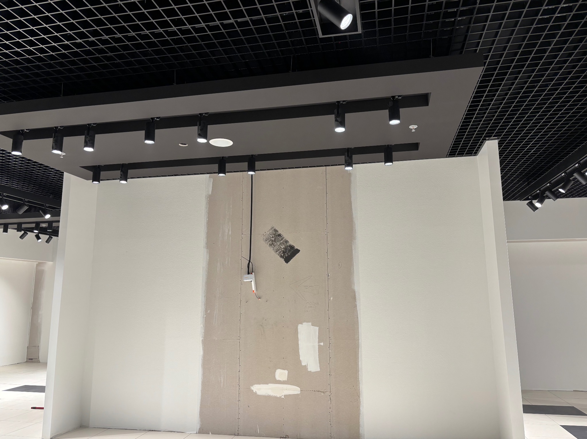 Ściana w trakcie szpachlowania w biurze, widoczne łączenia płyt karton-gips, instalacja elektryczna i czarna kratownica sufitowa z oświetleniem punktowym.