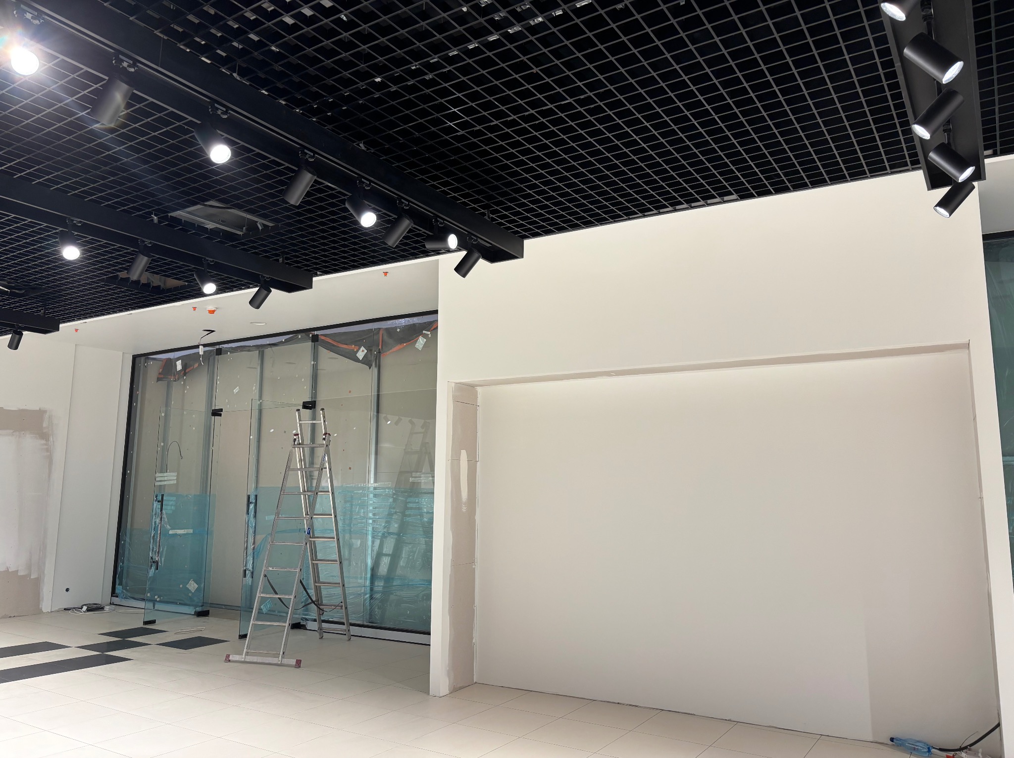 Wnętrze biura w trakcie wykańczania: białe ściany, aluminiowa drabina, przeszklona witryna zabezpieczona folią, czarna kratownica sufitowa z oświetleniem punktowym.