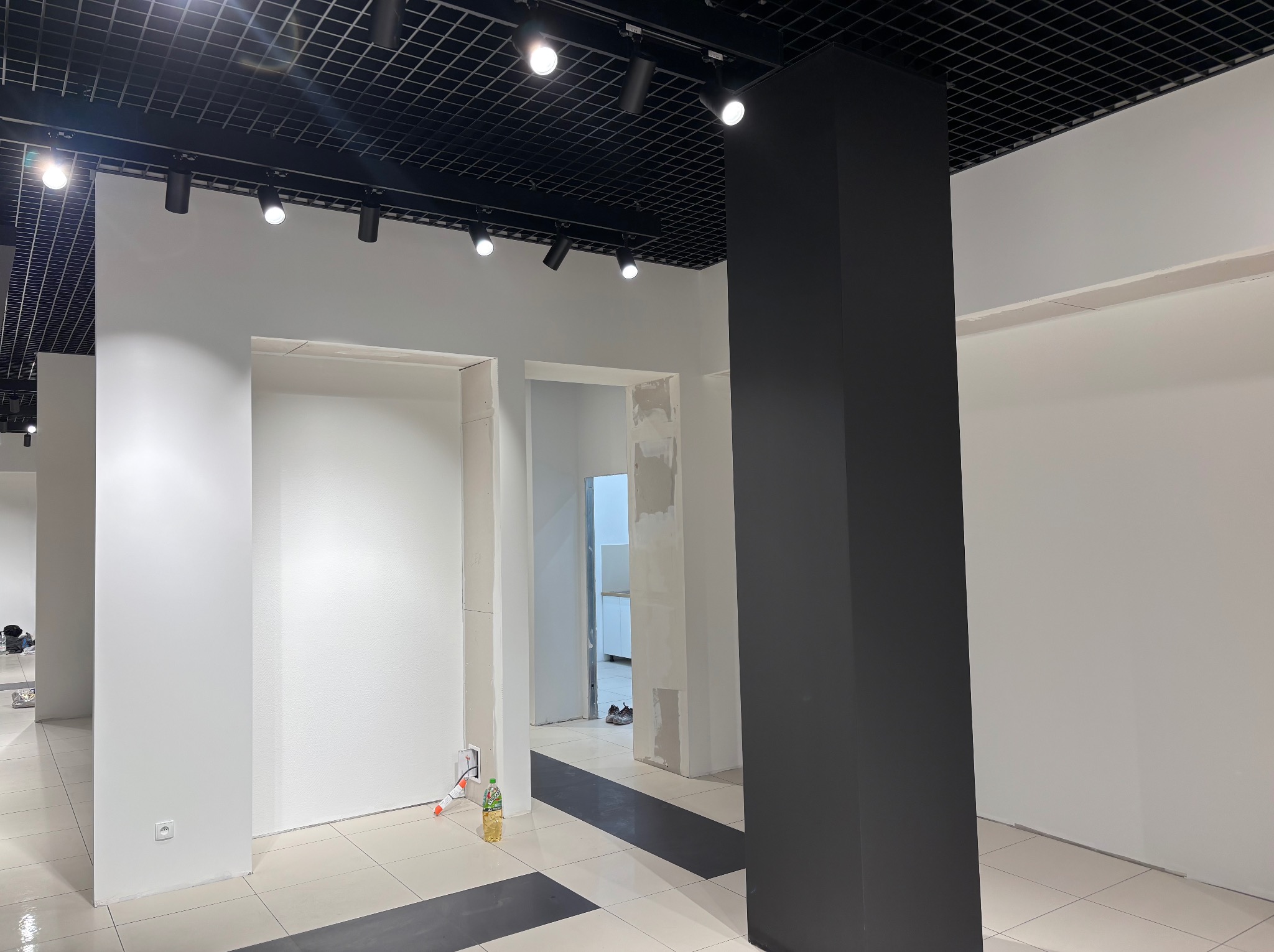 Wnętrze biura w trakcie wykańczania: białe ściany z otworami drzwiowymi, czarny filar, sufit z rastrem i oświetleniem. Widoczne ślady prac wykończeniowych.