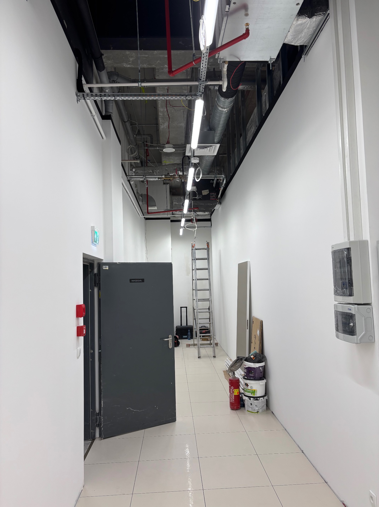 Korytarz biurowy w trakcie prac wykończeniowych: białe ściany, otwarta szara metalowa brama, drabina, puszki z farbą i instalacje pod sufitem.