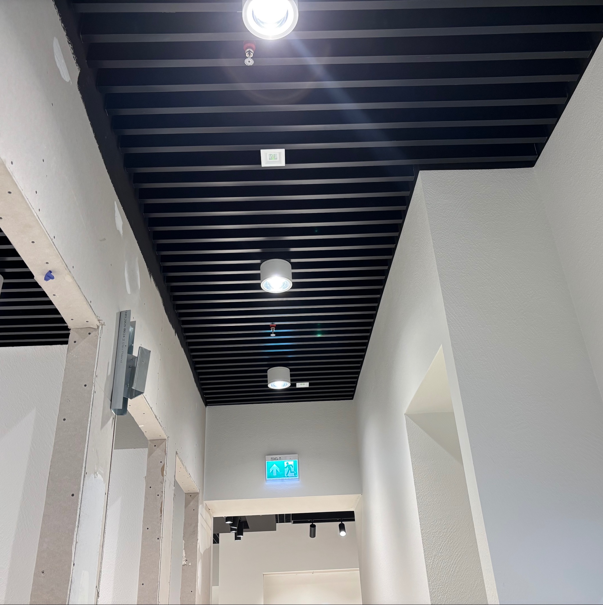 Sufit rastrowy w biurze z oświetleniem i czujnikiem dymu, ściany w trakcie wykańczania płytami gipsowo-kartonowymi, widoczne profile i elementy montażowe.