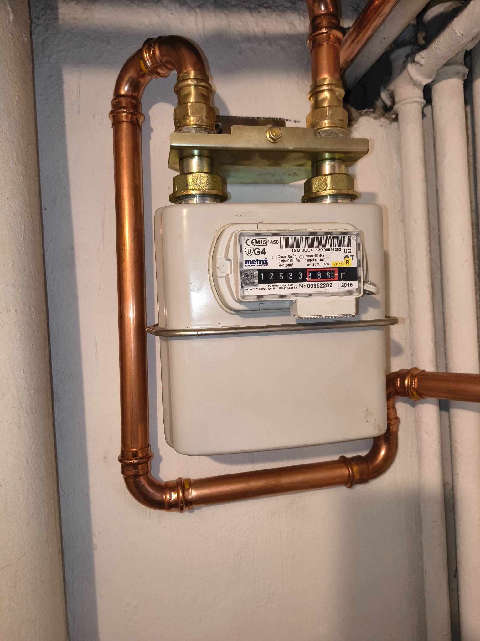 Zamontowany gazomierz z miedzianymi rurami i złotymi złączkami na białej ścianie. Widoczne numery seryjne i data produkcji na urządzeniu.