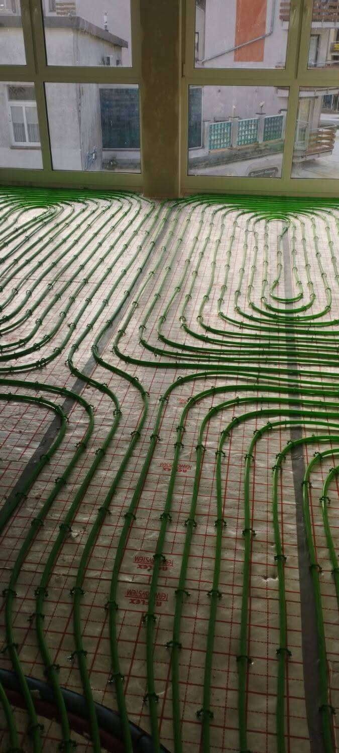 Zielone rury ogrzewania podłogowego ułożone na izolacji z siatką, widoczne przez okno budynku w tle. Instalacja przed wylaniem posadzki.