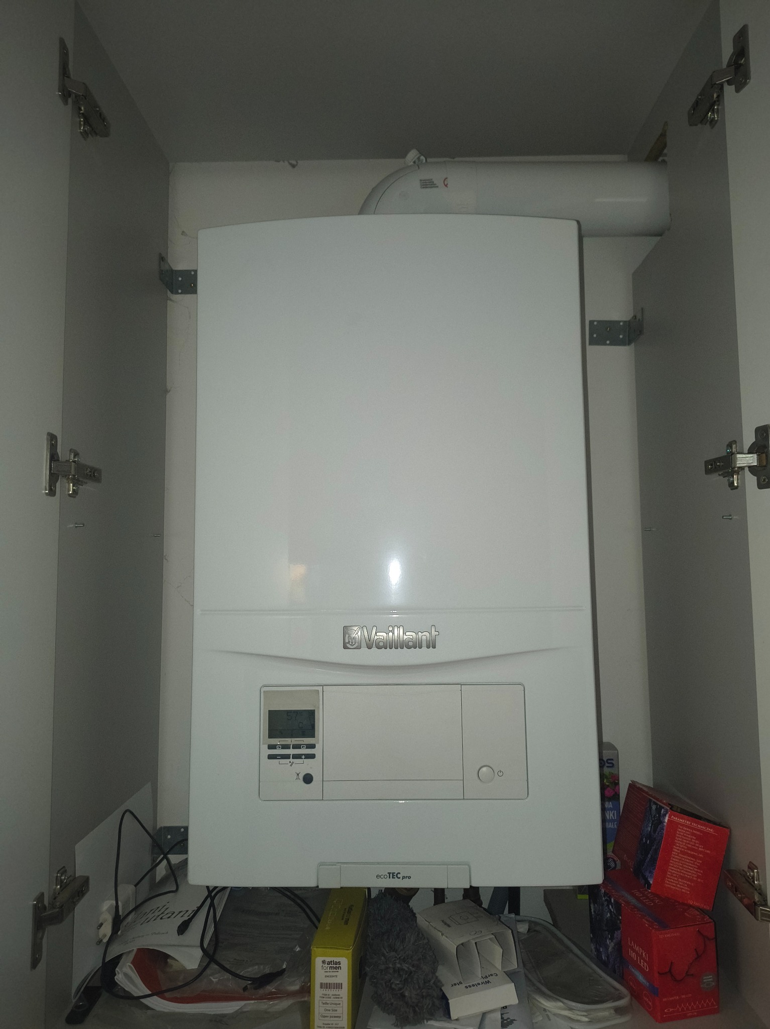 Biały piec gazowy Vaillant EcoTEC Pro zamontowany w szafce kuchennej, widoczne rury i elementy instalacji. Ujęcie z bliska, wnętrze szafki z drobnymi przedmiotami.