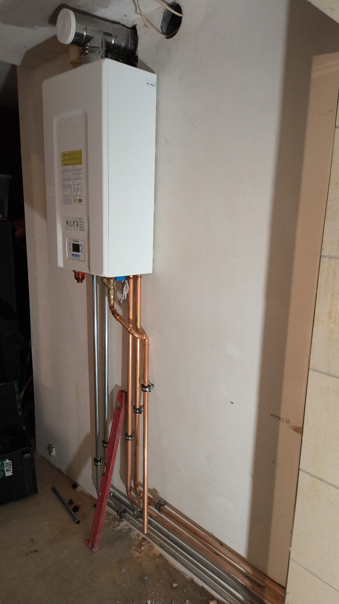 Biały piec gazowy Beretta zamontowany na ścianie z widocznymi miedzianymi rurami, poziomica i fragment podłogi z rurami. Instalacja w trakcie prac wykończeniowych.