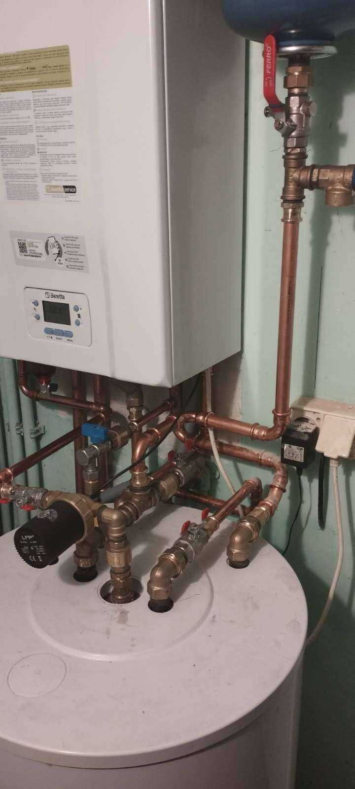 Biały piec gazowy Beretta z miedzianymi rurami i zaworami, zamontowany nad białym zasobnikiem wody. Widoczne przyłącza elektryczne i fragment ściany w tle.