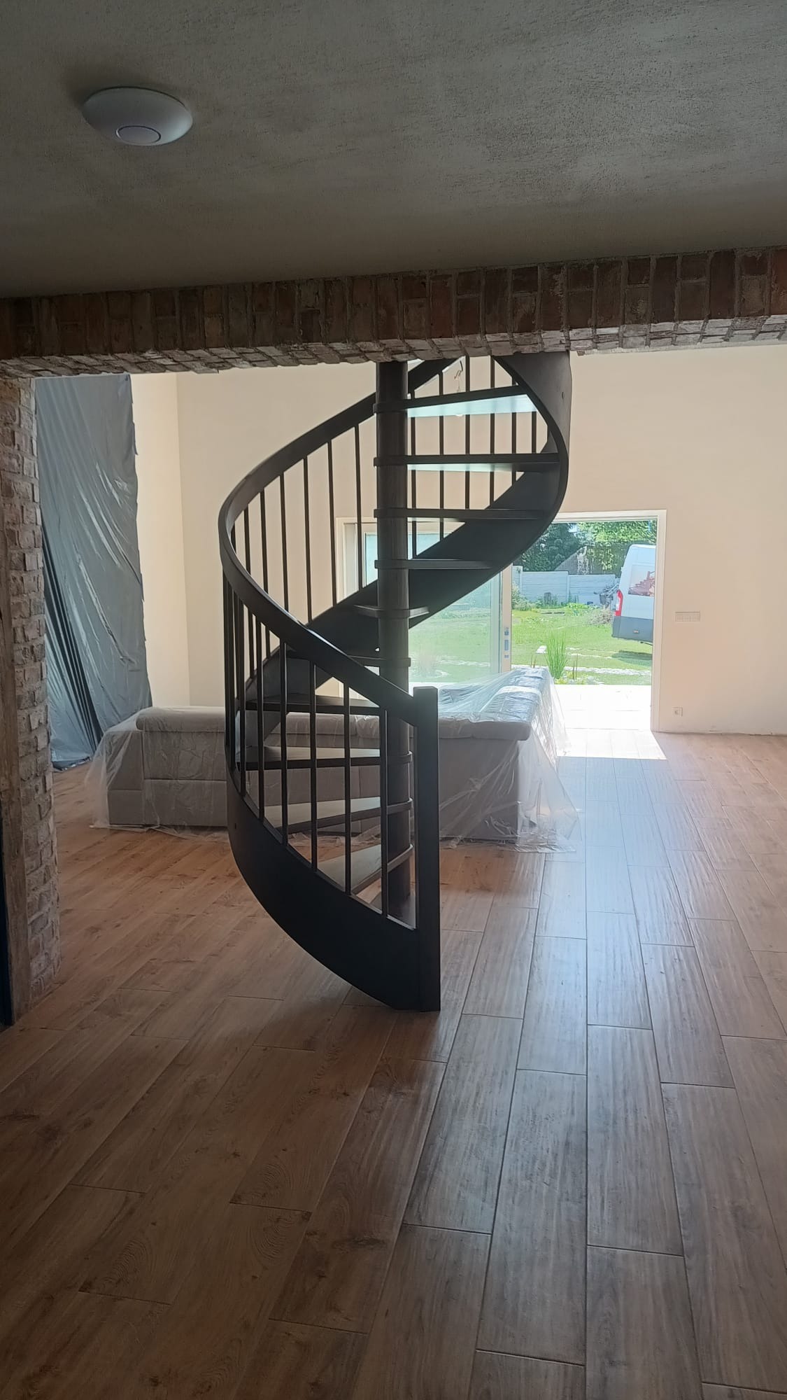 Spiralne schody metalowe w surowym wnętrzu z ceglanymi elementami i drewnianą podłogą. Sofa zabezpieczona folią malarską w tle.