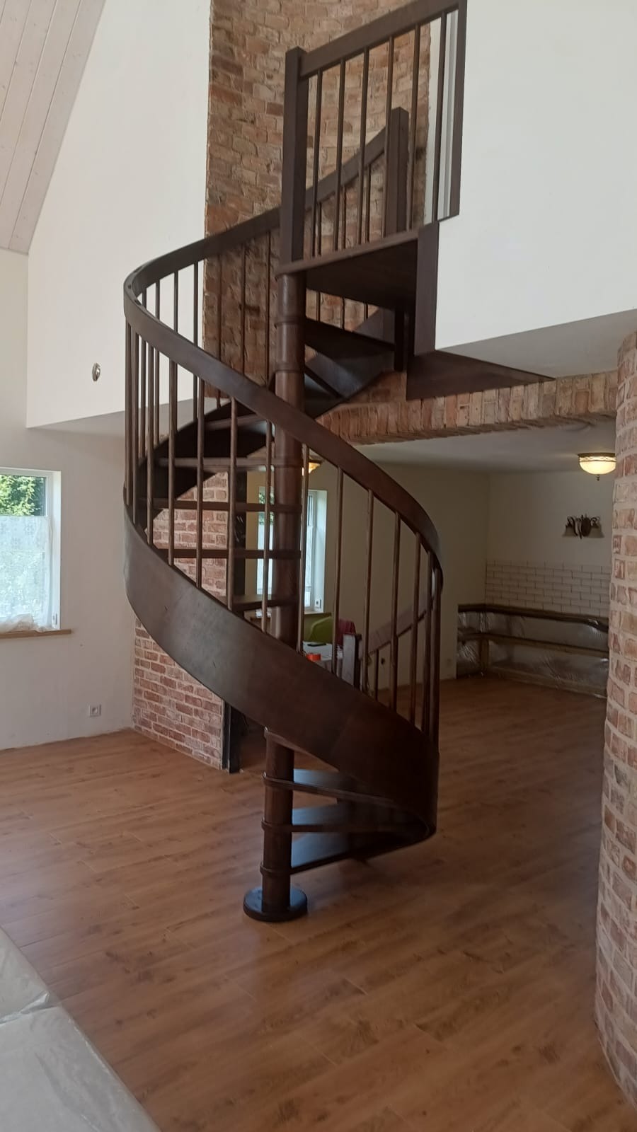 Spiralne schody drewniane w przestronnym wnętrzu z ceglanymi elementami, widoczne z perspektywy podłogi, ukazujące unikalny design i rzemiosło stolarskie.