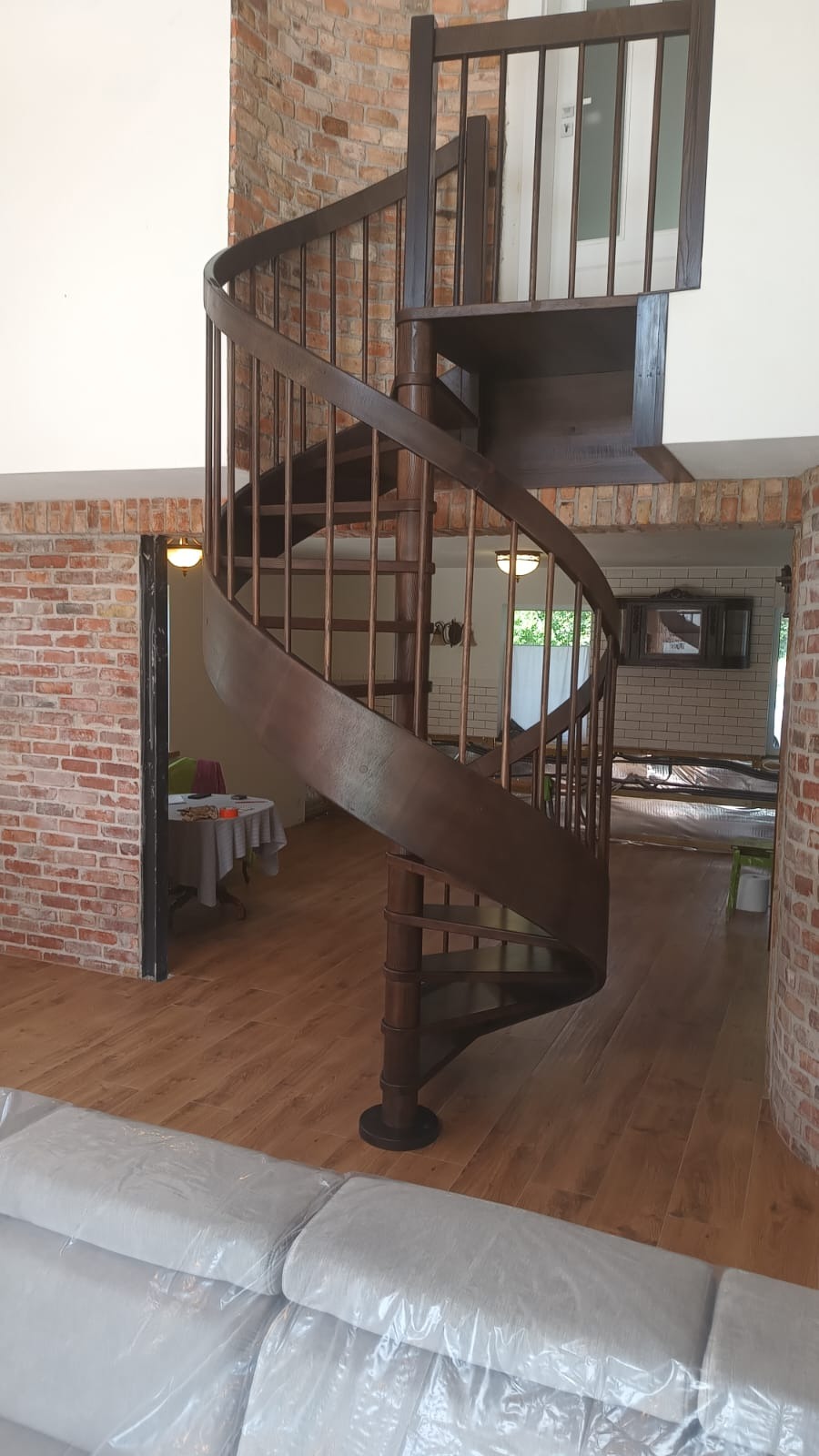 Spiralne schody drewniane w rustykalnym wnętrzu z ceglanymi ścianami i drewnianą podłogą. Widoczne zabezpieczone meble, sugerujące prace wykończeniowe.