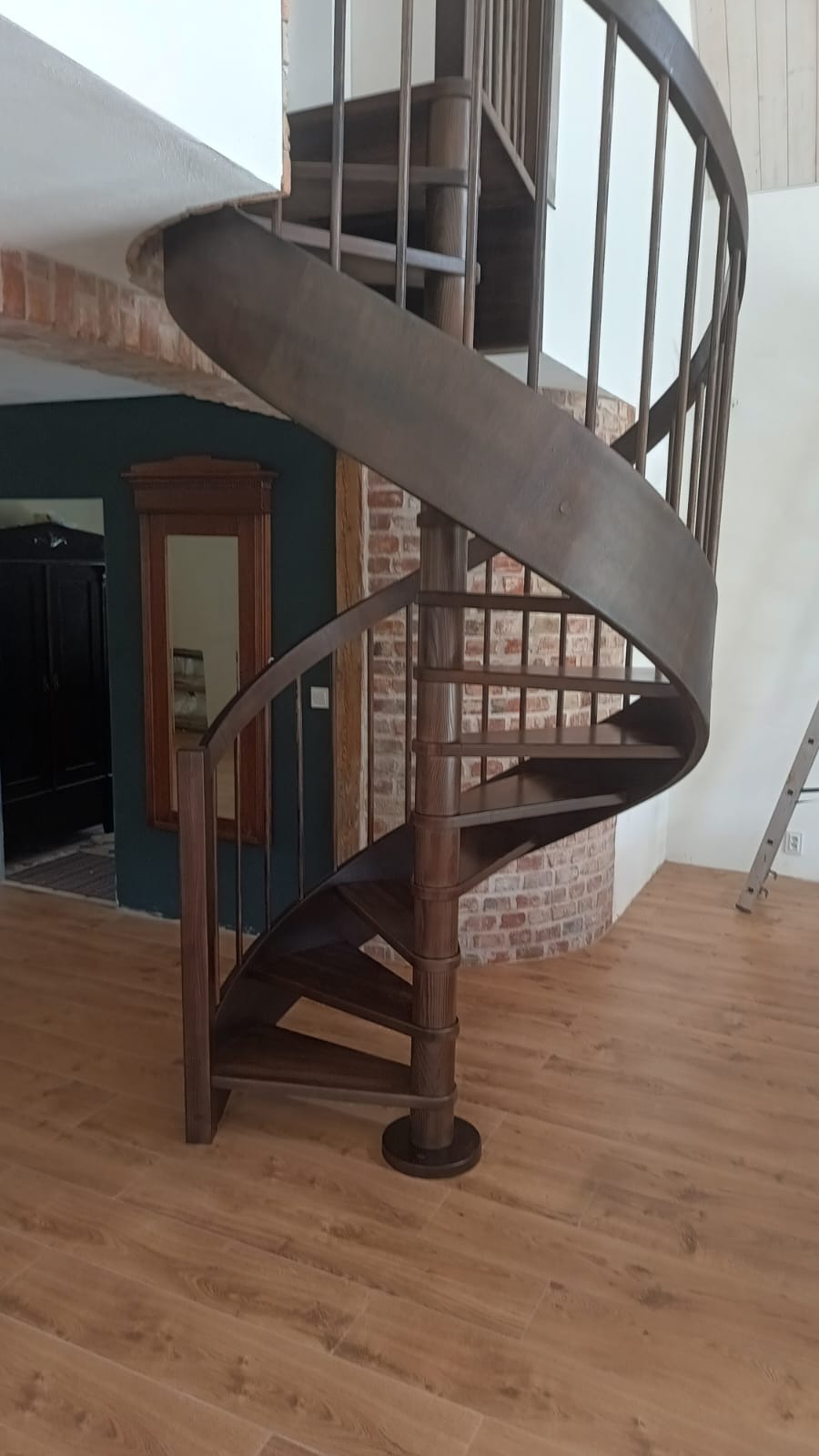 Drewniane schody spiralne w brązowym odcieniu, z metalową balustradą, na tle ceglanej ściany i drewnianej podłogi. Widoczne elementy wystroju wnętrza.