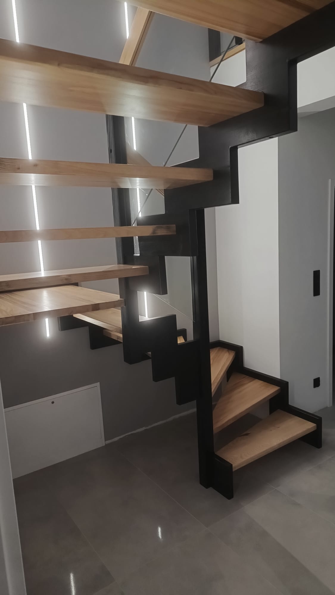 Nowoczesne schody drewniane z czarną metalową konstrukcją i subtelnym oświetleniem LED, widoczne z dołu, w minimalistycznym wnętrzu.