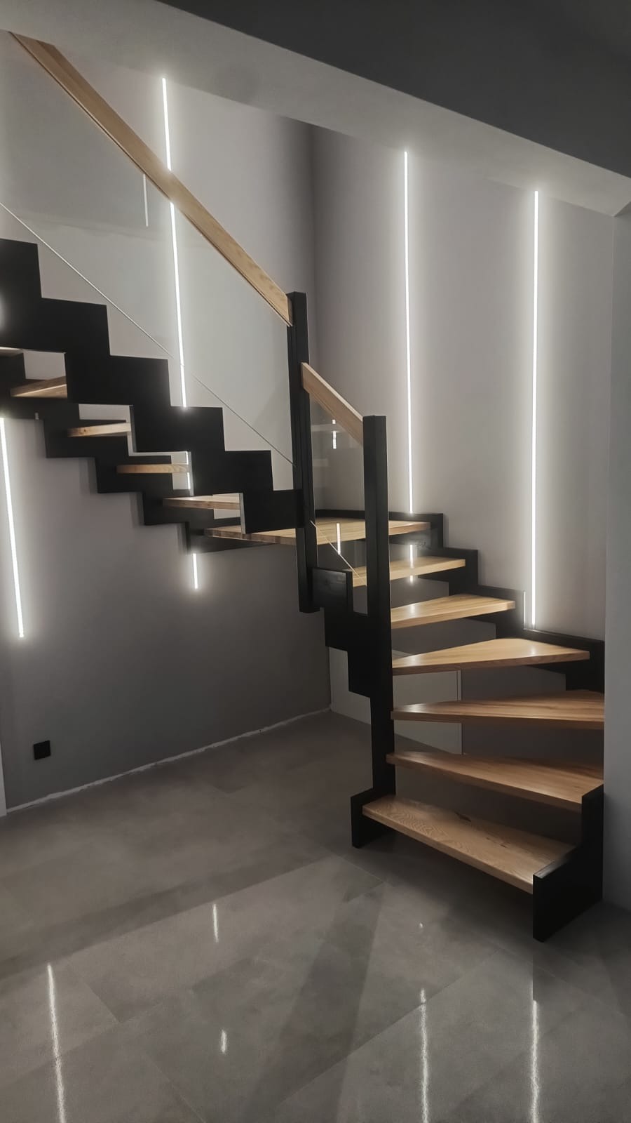 Nowoczesne schody drewniane z czarną konstrukcją i podświetleniem LED na ścianie. Jasne drewno stopni kontrastuje z ciemną ramą i szarą ścianą.
