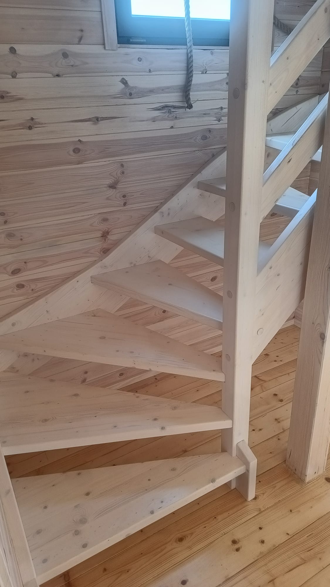 Jasne, drewniane schody z barierką w domu z boazerią, widoczne stopnie i fragment okna dachowego z liną. Naturalne oświetlenie podkreśla strukturę drewna.