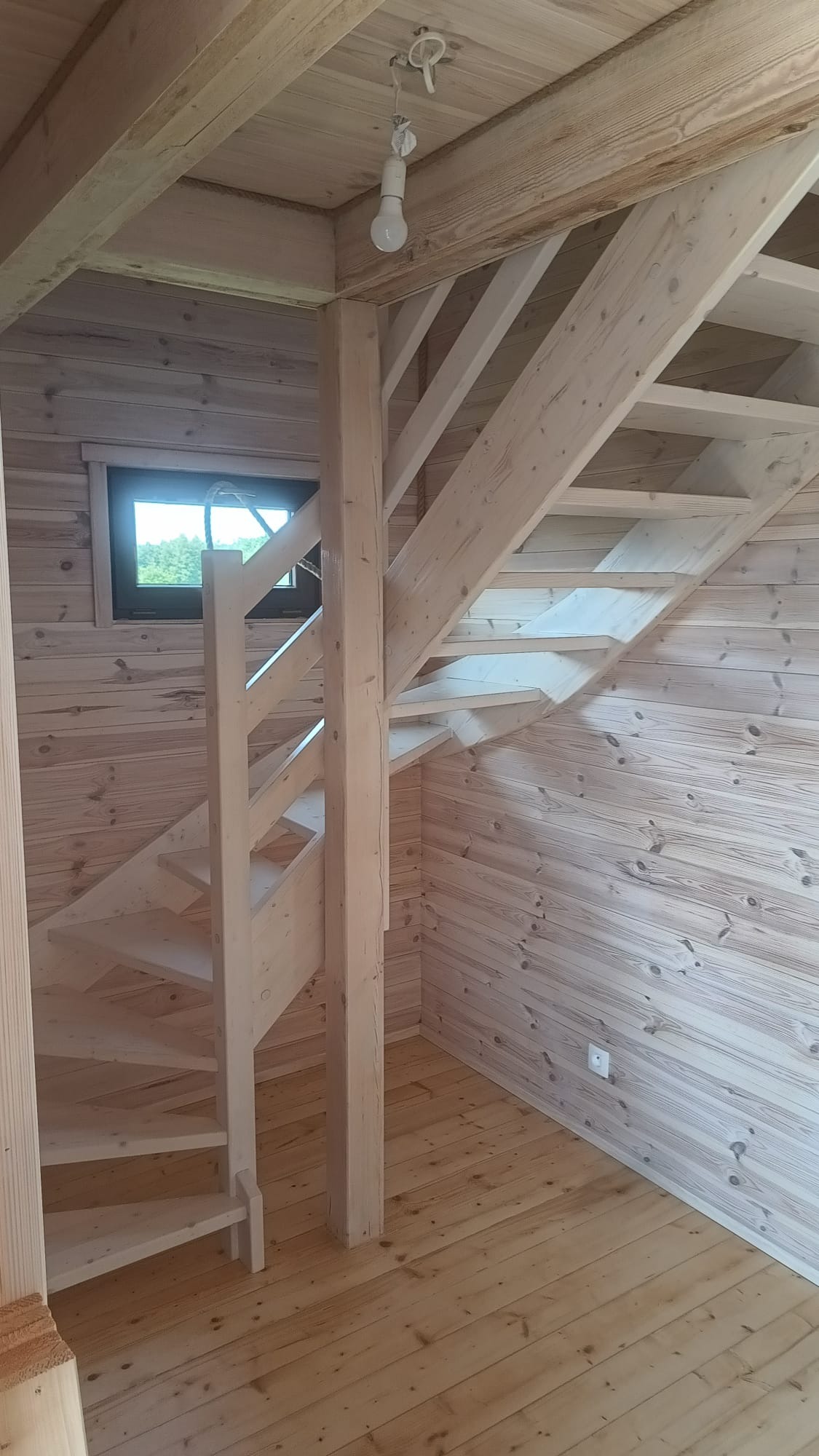 Jasne, drewniane schody spiralne w nowym domu z boazerią, oświetlone wiszącą żarówką. Widok z dołu na konstrukcję i małe okno w tle.