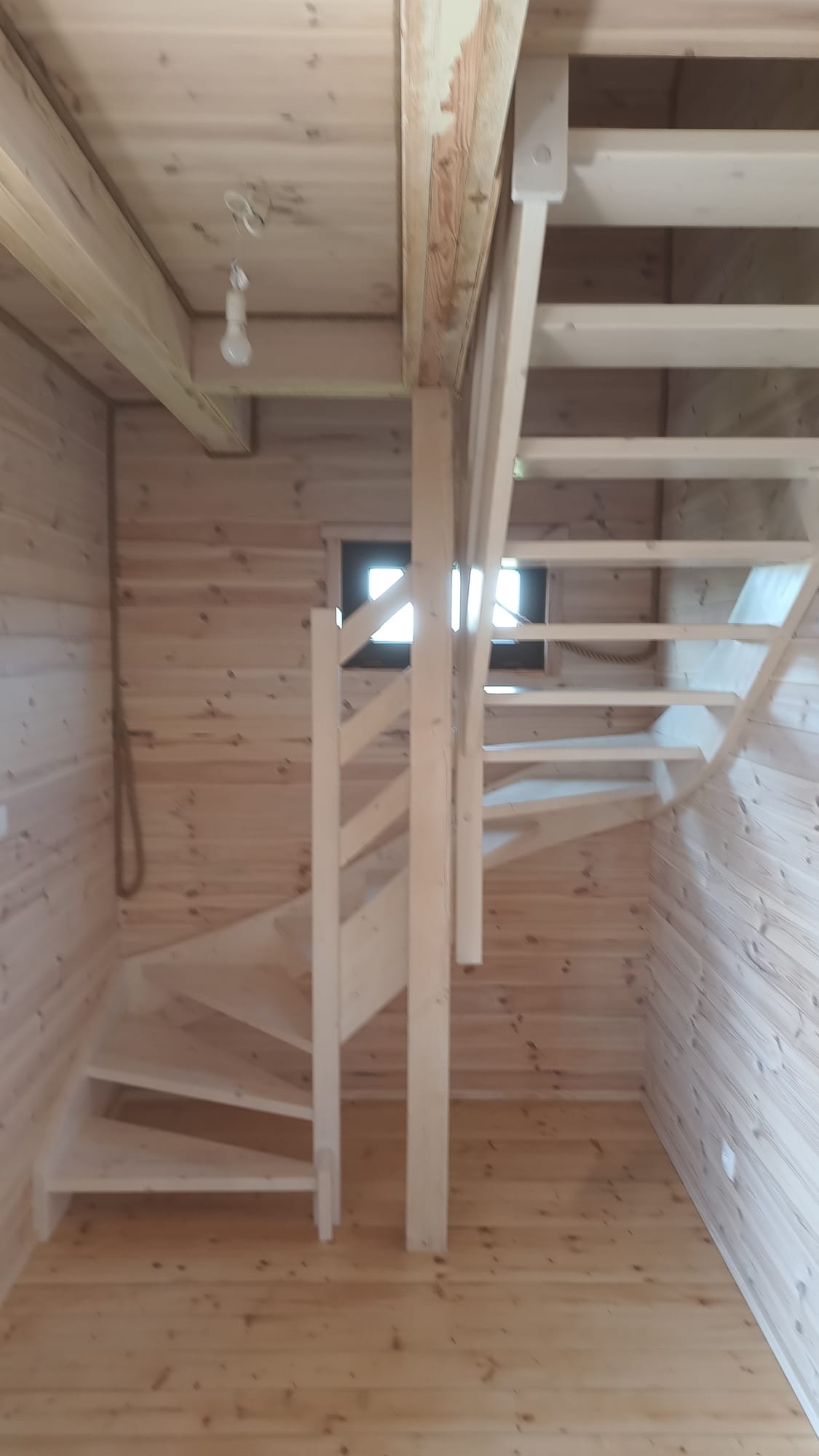 Drewniane schody spiralne w jasnym wnętrzu z boazerią, widoczne stopnie, poręcze i fragment okna. Naturalne oświetlenie podkreśla strukturę drewna.