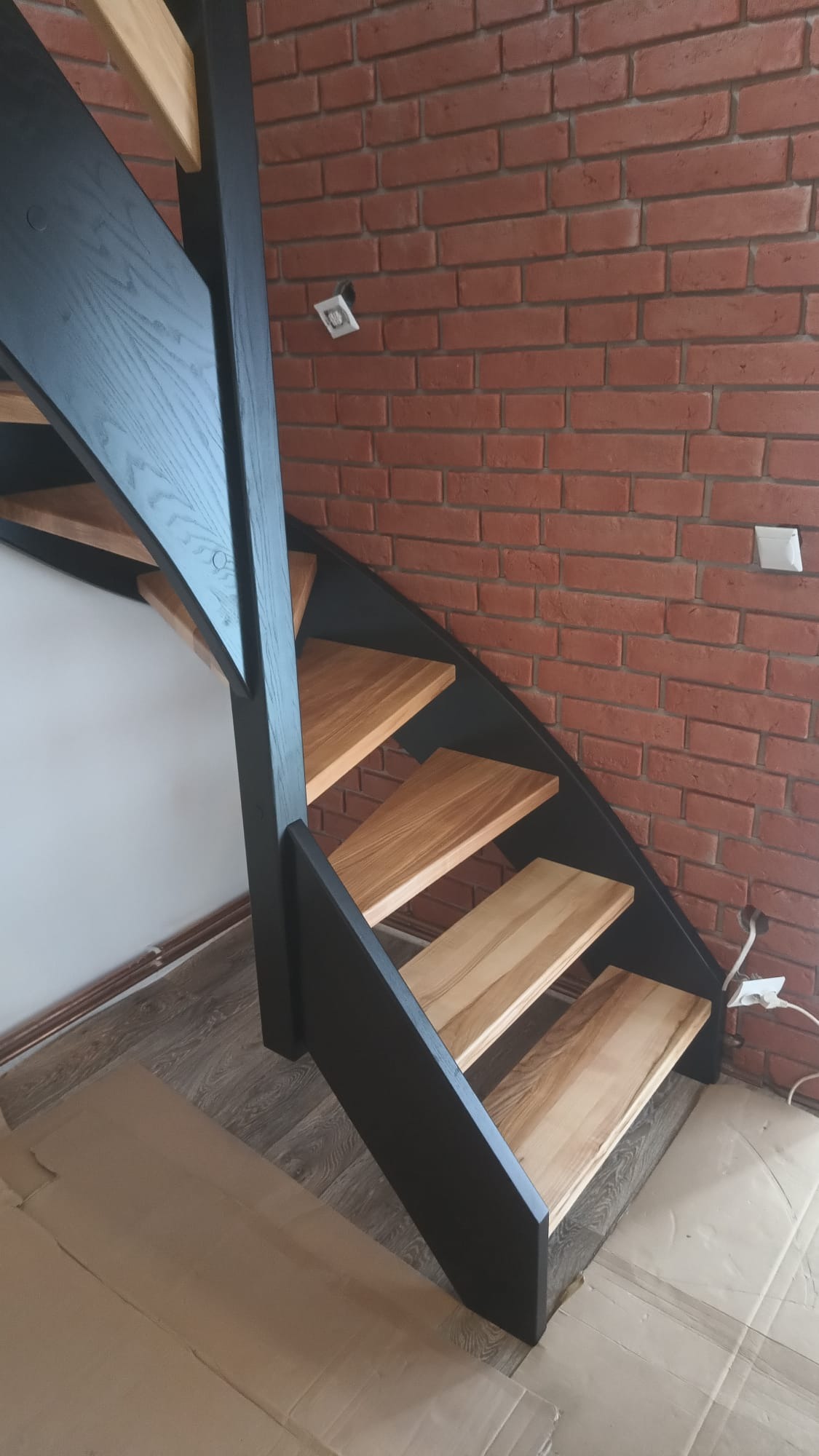 Nowoczesne schody drewniane z czarną balustradą i jasnymi stopniami, na tle ściany z czerwonej cegły. Widoczne gniazdka elektryczne i zabezpieczenie podłogi kartonem.