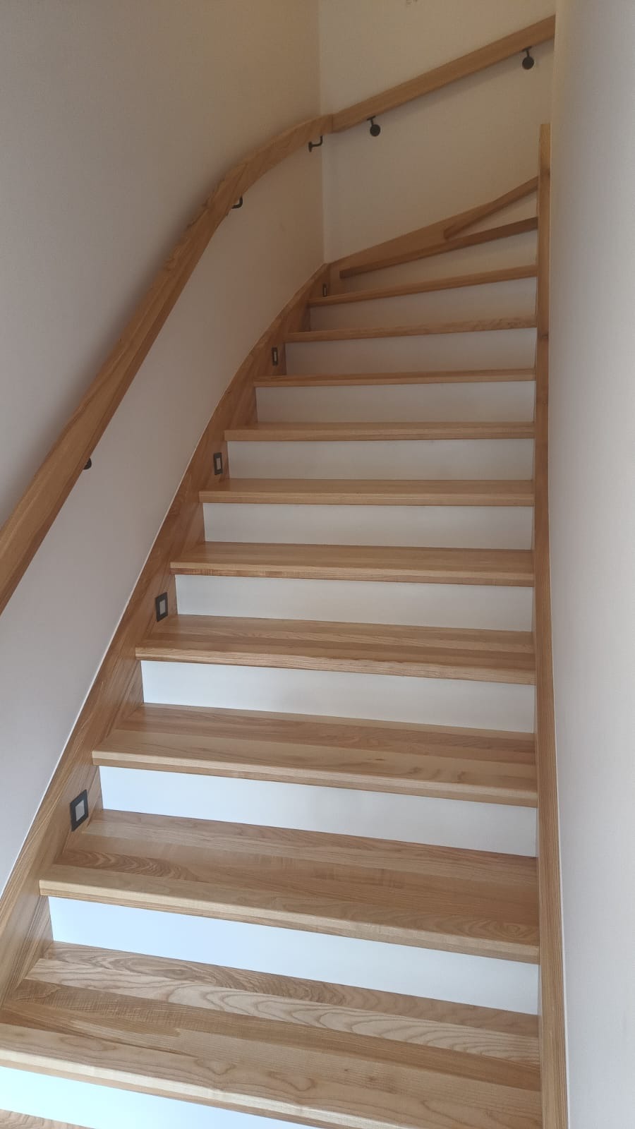 Jasne, drewniane schody z białymi podstopnicami, oświetleniem punktowym na ścianie, zakręcające w lewo, widok z dołu.