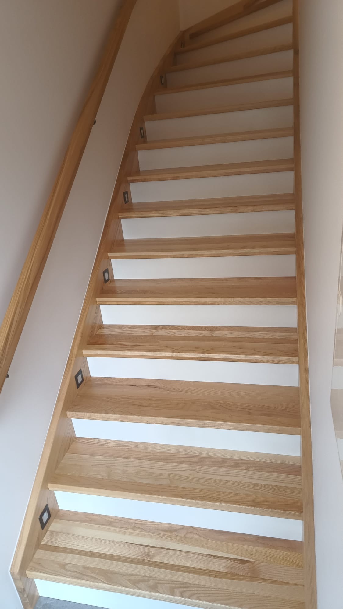 Zakrzywione schody drewniane z białymi podstopnicami i subtelnym oświetleniem punktowym w każdym stopniu, widok z dołu, jasne drewno.