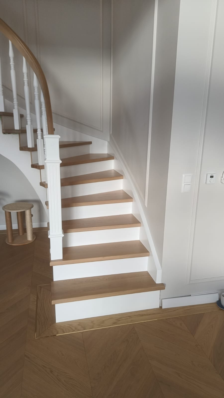 Drewniane schody z białymi podstopnicami i balustradą, parkiet w jodełkę, eleganckie wykończenie ścian w jasnym wnętrzu. Widok z dołu na pierwsze stopnie.
