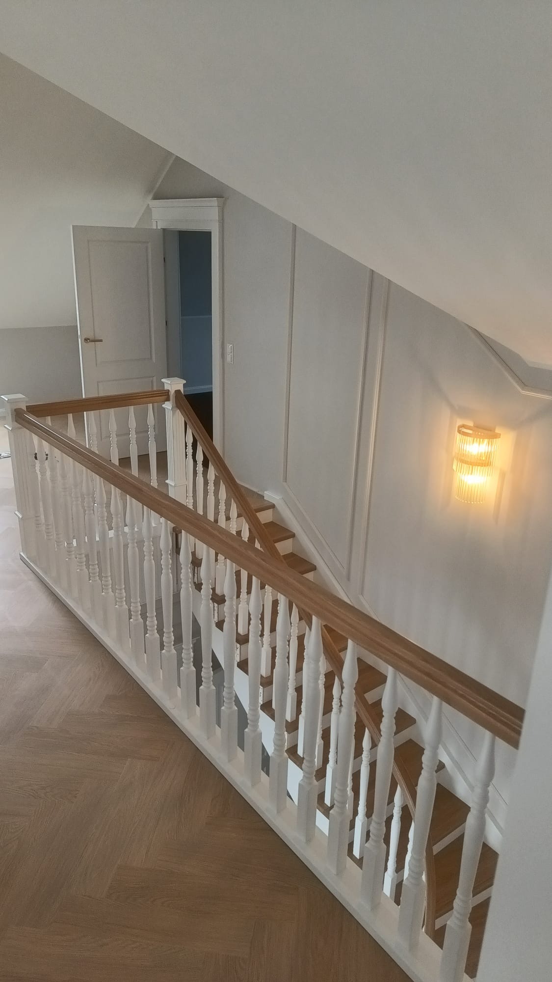 Jasne schody drewniane z białymi balustradami i drewnianymi poręczami, widok z góry, parkiet w jodełkę, oświetlenie ścienne, drzwi w tle.