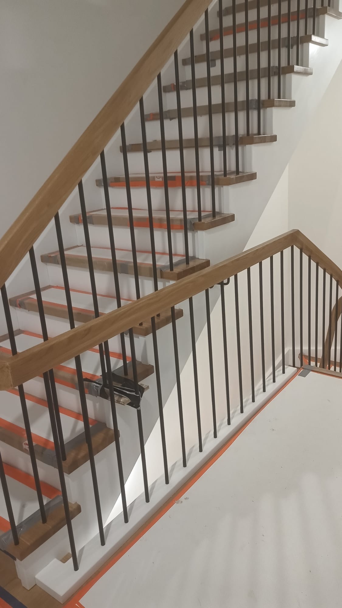 Nowoczesne schody z drewnianymi stopniami i czarnymi metalowymi balustradami, zabezpieczone folią ochronną podczas montażu. Jasne wnętrze, minimalistyczny design.